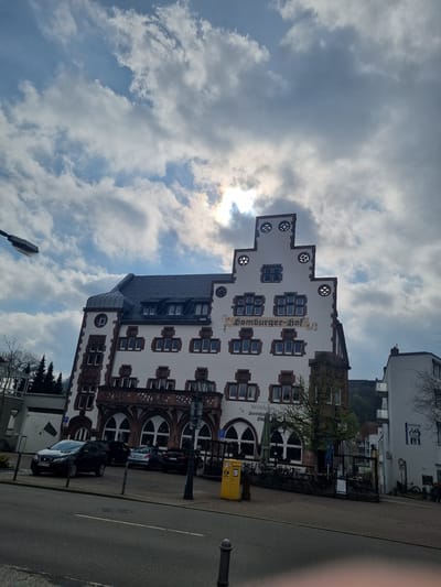 The image shows the Homburger Hof building in Homburg, Germany. The building is white with brown trim around the windows, and it has a distinctive stepped gable roof. The street alongside the building is lined with parked cars. The sky is cloudy, with the sun visible behind the clouds.