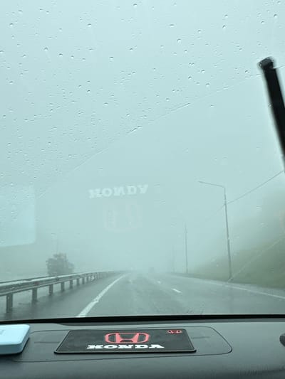 A car's windshield is covered in raindrops, obscuring the view of a foggy highway. In the distance, streetlights and a truck are barely visible. The dashboard features a Honda logo. The scene is the city of Svetly, Russia.