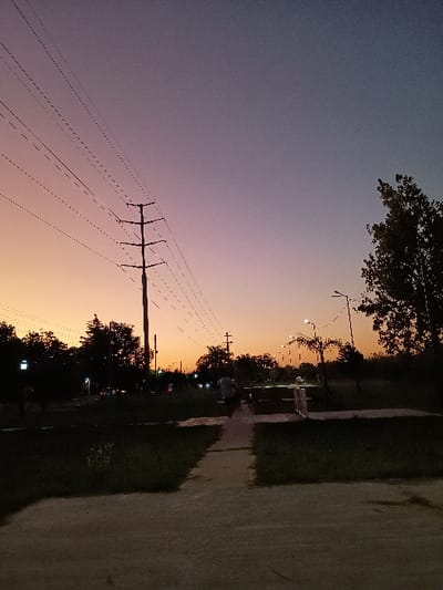 The image presents a serene outdoor scene captured during twilight in General Pacheco, Argentina. The sky dominates the upper portion, showcasing a dramatic gradient of colors, transitioning from a deep blue-purple at the top to vibrant orange and yellow hues as it approaches the horizon.
In the left foreground, a prominent utility pole stands tall, extending upwards and featuring multiple dark power lines stretching across the colorful sky. On the right side of the frame, a concrete path or sidewalk runs horizontally, bordered by dark, grassy areas and silhouetted foliage, suggesting a park or suburban setting.
Along the distant horizon, dark, indistinct shapes of trees are silhouetted against the bright sky. Several small, bright points of light, likely streetlights or distant vehicle lights, are visible further along the path in the background. The lighting suggests either sunrise or sunset, creating a peaceful and contemplative atmosphere. There are no clearly discernible people or specific activities taking place, and no visible text is present.