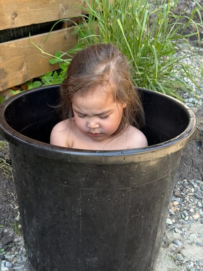The image depicts a young child, appearing to be a toddler, situated within a large, black, plastic utility tub outdoors in Kyzyl-Ozek, Russia. The child is partially submerged in water, visible from the upper chest, and appears unclothed. The child has light brown hair, which shows signs of moisture, and is looking downwards with a focused expression, presenting furrowed eyebrows.

The black plastic tub is cylindrical, exhibiting water droplets and streaks on its exterior surface, indicative of its current use. The immediate ground surrounding the tub is composed of a mixture of dark soil, small pebbles, and larger stones. In the background to the left, a wooden structure, likely a fence or wall, features horizontal planks of weathered dark brown wood. Green foliage is visible beneath and between these planks. To the right background, a dense growth of tall, green grass or reeds is present, behind which the ground consists of mixed gravel and dirt. The scene is illuminated by natural daylight.