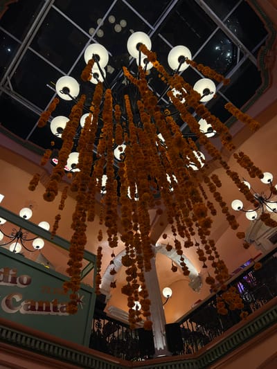 This image captures the interior of a multi-level building in Guanajuato City, Mexico, specifically an area associated with "Radio Canta 77.7 FM," as indicated by a prominent sign on an elevated green panel to the left.

The central focus is a large, ornate black metal chandelier suspended from a glass-paned ceiling or skylight. The chandelier features numerous spherical white light fixtures and is extensively adorned with cascading strands of bright orange marigold flowers, varying in length and creating a dense, floral curtain effect. These marigold decorations are consistent with Día de Muertos cultural observances.

The surrounding architecture features pale orange/peach-colored walls, vaulted ceilings, and white stone columns and arches. Dark, ornate metal railings delineate at least two visible levels of the interior space. Additional smaller chandeliers or wall-mounted sconces with spherical white lights are visible throughout the lower and upper levels. Two dark, rectangular speakers are positioned near the railings on a lower level. No individuals are present in the scene. The overall lighting is artificial and warm, emanating from the various fixtures, contrasting with the dark exterior visible through the glass ceiling.