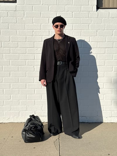 A person stands in front of a white brick wall wearing a black beret, sunglasses, a dark striped shirt, a black blazer with a small cross pin, and black pants with a black belt. The person is also wearing black shoes. A black leather bag sits next to the person. The sun casts a shadow of the person and the top of the wall.