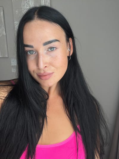 A medium shot shows a young woman with dark hair and fair skin with freckles. She is wearing a bright pink top. Her eyes are light blue, and she has dark eyebrows. She is wearing a small hoop earring in her left ear. The background appears to be a room with grey walls and a curtain. There is a framed artwork on the wall behind her. The image is a selfie, taken from a slightly high angle. The location is Kokkorevo, Russia.