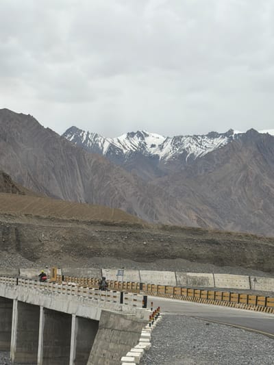 A motorcyclist wearing a yellow jacket and helmet rides on a paved road with a concrete bridge. The bridge has a yellow and black striped guardrail and concrete supports. In the background, a mountain range with snow-capped peaks is visible under a cloudy sky. The surrounding terrain is rocky and barren. The location is Zangla, India.