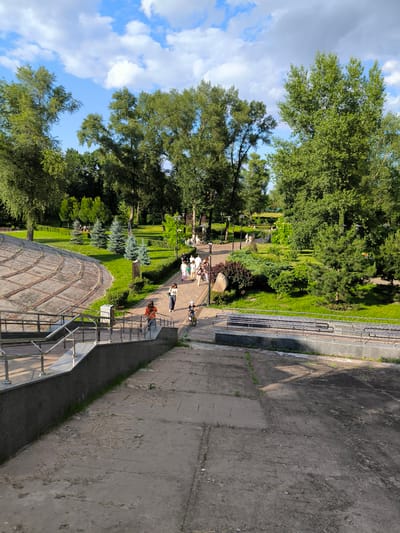 The image depicts a park scene with a concrete pathway and stairs in the foreground, leading up to a green lawn with scattered trees and people walking. The park is surrounded by lush green trees and under a partly cloudy blue sky. In the middle ground, a group of people is walking along a path, some appear to be a family with a child on a bicycle. The pathway is lined with benches and lampposts. In the background, more trees and foliage are visible, creating a serene park atmosphere. The overall scene suggests a pleasant day outdoors in a public park.