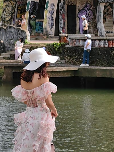 A woman with auburn hair, wearing a white wide-brimmed sun hat and an off-the-shoulder white dress with a pink floral pattern and ruffled tiers, is depicted from behind, facing a body of green water. She holds a light-colored bag in her right hand. The scene is located in Kim Long Ward, Vietnam.
Behind the water and the woman, a multi-level concrete structure with a dark, textured base is visible. This structure is heavily adorned with various colorful graffiti, including abstract shapes, stylized letters, and a large green and brown face with a prominent eye. Several individuals are present on and around this structure:
* On an upper level to the left, a woman with blonde hair, wearing a light-colored top and skirt, stands near a wall.
* Below her, on a set of concrete steps, another woman in a light top and striped pants sits.
* Further down and to the right, a person in a white bucket hat and a white top is seated on a lower ledge.
* On a more prominent platform to the right, a woman in a white t-shirt, blue jeans, white sneakers, and a white baseball cap stands facing to the left, holding a small dark bag.
* Above her, on a higher section of the structure, another woman in a white top and cap is partially visible, appearing to walk towards the right.
The overall environment suggests an outdoor recreational area with a distinctive architectural element featuring street art, bordering a calm body of water under bright daylight.