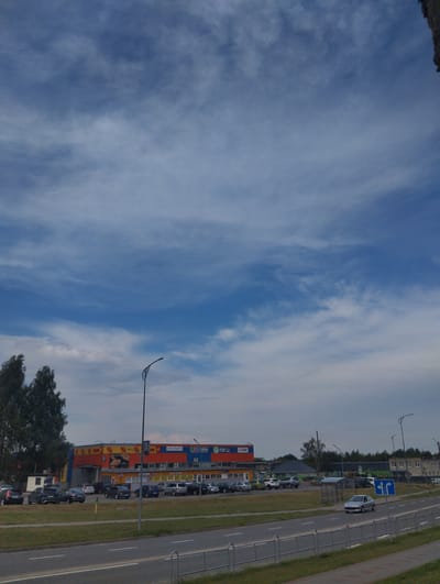 A wide shot captures a suburban street scene with a prominent commercial building. The sky is a vast expanse of blue with wispy white clouds, suggesting a daytime setting with fair weather. On the left, a cluster of dark green trees stands tall.

In the foreground, a paved road with lane markings is visible, bordered by a grassy median and a grey metal fence. Several cars are parked in front of a long, colorful building. The building features a bright orange and yellow facade with a red section and a blue awning. Visible text on the building includes "FIXPrice" and "TOR" in red and blue, along with various logos and advertisements, some depicting pizza slices. To the right of the commercial building, a modern streetlamp with a curved arm stands tall.

Further down the road, more cars are parked, and other buildings, some residential with light-colored facades and windows, can be seen in the distance. Another streetlamp is visible further along the road. A blue directional sign with arrows is placed near the right edge of the road, indicating lane choices. The overall scene depicts a commercial area on a clear day.