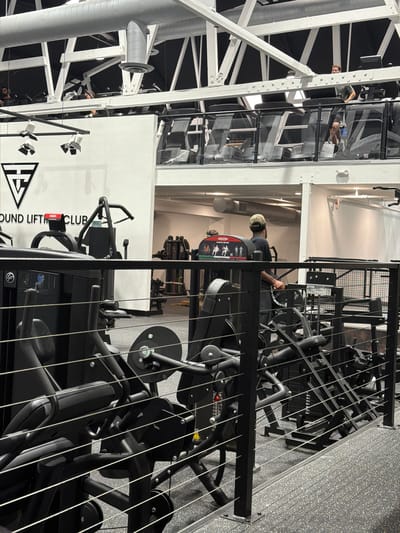This indoor shot depicts the interior of a gym, identified by the signage "GROUND LIFTING CLUB" and a logo on a white wall, presumably located in Solana Beach, United States of America. The gymnasium is equipped with weightlifting machines. The machines are primarily black. A person is seated at one, with a camouflage cap and dark clothing. Another person is visible on a raised platform in the background. The gym features a complex network of white structural supports and piping on the ceiling. The presence of a second-story mezzanine is noted. The flooring is speckled gray. The overall lighting in the gym is bright.