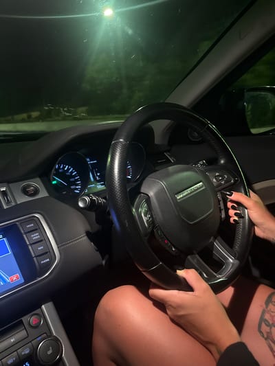 The image provides an interior view of a vehicle at night, likely in the Metropolitan Borough of Solihull, United Kingdom. The central focus is a person's hands gripping the black leather steering wheel of a Range Rover, whose emblem is prominently displayed. The left hand, adorned with black nail polish, is positioned on the top left quadrant of the steering wheel, near control buttons, while the right hand rests on the lower right.
A partial view of the person's upper right leg is visible, revealing a tattoo. The vehicle's dashboard is illuminated, with the instrument cluster showing a speedometer indicating approximately 5 mph (and km/h markings). A "START STOP" button is located below the cluster. The center console features an infotainment screen, partially displaying a map or navigation interface with a red "X" icon. Surrounding buttons are labeled "MODE", "AUDIO", "VIDEO", "NAV", and "PHONE". To the right, climate control dials, one marked "TEMP", and a hazard light button are visible.
The scene is dark, suggesting nighttime, with external lights reflecting on the windshield and rearview mirror.
Visible text includes: "RANGE ROVER" on the steering wheel, "START STOP" on a button, "MODE", "AUDIO", "VIDEO", "NAV", "PHONE" on console buttons, and "mph" and "km/h" on the speedometer. Numbers like 20, 40, 60, 80, 100, 120, 140, 160 are also visible on the speedometer.