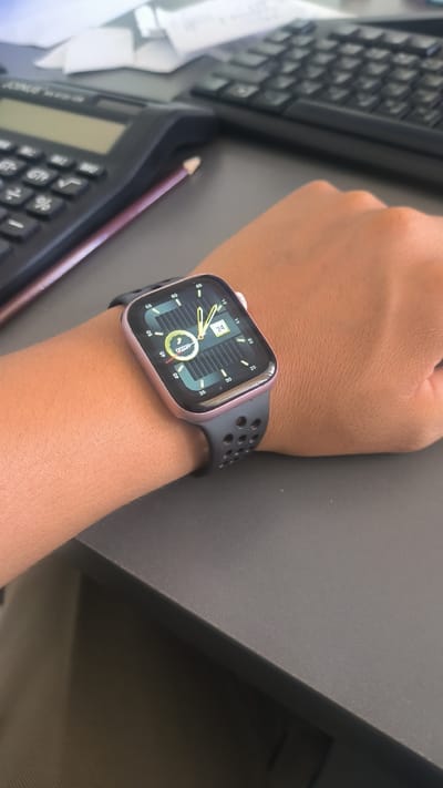 A close-up, eye-level shot shows a person's left arm wearing a rose gold Apple Watch with a black perforated sport band. The watch face displays a digital readout of "24" within a square, and two analog hands, one indicating approximately 3:30. A circular graphic with a yellow center and segmented green and black outer rings is also visible on the watch face. The watch is resting on a dark grey desk, where parts of a black calculator and a black computer keyboard are visible in the background. A pink pencil is positioned between the calculator and the watch. The overall lighting suggests an indoor setting, possibly an office or workspace. The image is cropped to focus on the watch and arm, with the background elements slightly blurred. No specific location in Булак-Башы, Kyrgyzstan is discernible from the image.