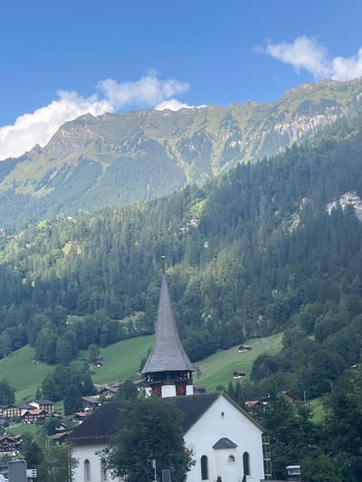 An outdoor, daylight image captures the Lauterbrunnen Reformed Church in Lauterbrunnen, Switzerland, positioned in a valley. The church features white walls, dark roofs, and a prominent, tall, conical steeple with dark grey shingles, accented by a band of red and white decorative elements below the spire. The base of the church is partially obscured by trees and other structures, indicating part of the village. Behind the church, steep mountain slopes rise abruptly, densely covered with coniferous forests in the mid-ground. Above the tree line, the upper mountain sections display exposed rock, grassy areas, and visible terracing. A small building or structure is discernible on a distant mountain ridge. The sky above is clear blue with scattered white cumulus clouds. No individuals are visible.