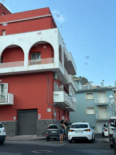 The image shows a three-story building in Giardini-Naxos, Italy, with a red facade and white arched balconies. A gray garage door is at street level. Adjacent to the building is a light blue building with balconies. Several parked cars line the street, and people are visible next to one of the white cars. The sky is blue with a few clouds.