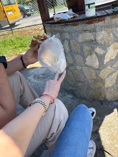 The image presents an overhead shot of two individuals, likely seated outside, sharing a meal. One person is holding a sandwich wrapped in a clear plastic bag. Their hand is adorned with a black and red beaded bracelet. On the same wrist, a black smartwatch is visible. The other person's hand is making a peace sign, and they have a red string bracelet on their wrist. A silver-colored watch is on their forearm. The individuals are wearing casual clothing, jeans and pants. The setting appears to be a roadside in Probuda, Bulgaria, indicated by the presence of a fence, a yellow van, and greenery in the background. The van's license plate reads TX-01-89-CT, and the surrounding buildings appear residential. The surface they are sitting on is made of stone.