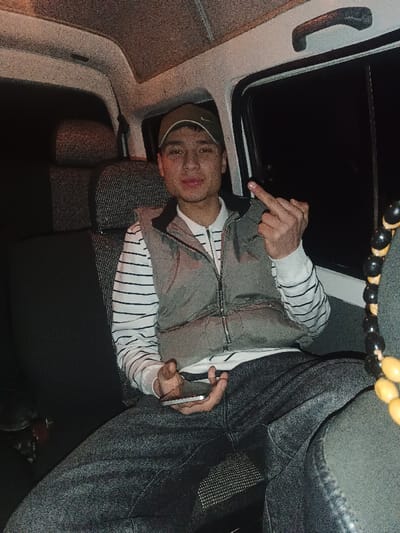 The image captures a young man seated in what appears to be the back seat of a vehicle, likely a van or a car with a high roof. The setting suggests it is nighttime or very dark outside, as the windows show only blackness.
The man, who appears to be in his late teens or early twenties, is positioned centrally, looking directly at the camera. He has a light skin tone, dark eyes, and a slight, almost smirk-like expression on his face. He is wearing a dark baseball cap, possibly olive green or dark grey, with a lighter-colored logo on the front that resembles a Nike swoosh. Over a long-sleeved white shirt with horizontal black or dark stripes, he wears a grey puffer vest with a black collar and a visible zipper down the front. He is also wearing dark jeans, possibly grey or black, which appear somewhat faded.
His left hand (viewer's right) is holding a silver or white smartphone. His right hand (viewer's left) is raised, with the middle finger extended in a clear "fuck you" gesture.
The interior of the vehicle is visible. The seats are upholstered in a dark, textured fabric, likely black or dark grey, with a visible patterned weave on the backrests. The ceiling is light-colored and shows some texture. A grab handle is visible above the window on the right side. To the right of the man, hanging from the ceiling or a fixture, is a strand of large, round beads, alternating in color between black and yellow or light brown.
The overall lighting is dim, suggesting low-light conditions, and the image has a grainy quality, particularly noticeable on the man's skin and in the darker areas of the background. No discernible text is visible in the image.