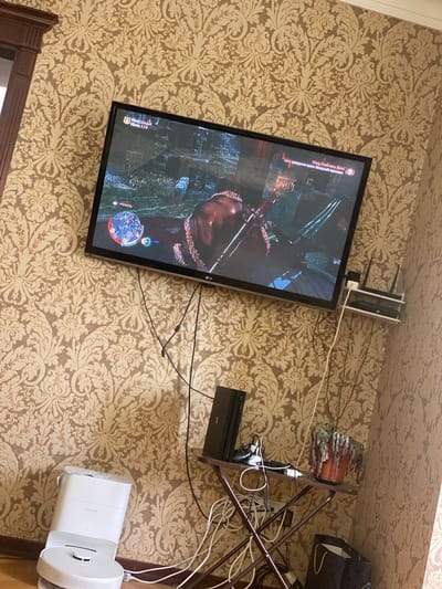 The media file depicts a television screen displaying a video game, likely an action or adventure game with a fantasy theme, given the character's attire and the in-game map. The television is mounted on a wall covered with ornate, floral-patterned wallpaper in beige and brown tones.
Several electronic devices are visible around the television, including what appears to be a game console or media player connected to the TV via cables. Some cables are neatly organized, while others are tangled, suggesting a functional but not perfectly managed setup. A white electronic device, possibly a smart home speaker or router, is positioned on the floor near the bottom right of the image.
On a shelf above and to the right of the television, there is a decorative item resembling a stylized potted plant with a reddish-brown trunk and layered, leaf-like elements. The overall setting suggests an indoor environment, likely a living room or entertainment area in a residential space. The lighting appears to be artificial, as is common for indoor spaces where televisions are used. There are no obvious cues indicating the time of day or weather conditions.
The text visible on the television screen is in Russian. One prominent piece of text reads "Могу любить Воля" which translates to "I can love Will". Another phrase "Убийство 1/4" translates to "Kill 1/4", indicating progress within the game. The setting is specified as Bogatyryovka, Russia.
