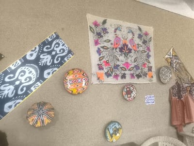 On a textured, light brown wall in an indoor setting in Turkkishlak, Uzbekistan, various traditional Uzbek crafts are displayed. A prominent square, translucent textile in the upper center is intricately embroidered with two confronting peacocks, stylized floral motifs, and numerous sequins. To its left, a long, diagonally oriented fabric strip features dark blue and white Ikat-style patterns. Below the central textile and to its left, multiple round, domed objects with vibrant geometric patterns, resembling skullcaps or decorative plates, are mounted. On the upper right, another rectangular textile with geometric patterns is visible, along with several draped garments or fabrics adorned with sequin embellishments hanging below it. A small, circular framed artwork depicting a building or landscape scene is present in the lower center. To its right, a white rectangular sign with blue text reading "SUBHAN SELEC UZBEK" is affixed. A fragment of white wicker furniture is visible in the bottom right corner of the frame. No people, actions, or direct interactions are depicted.
