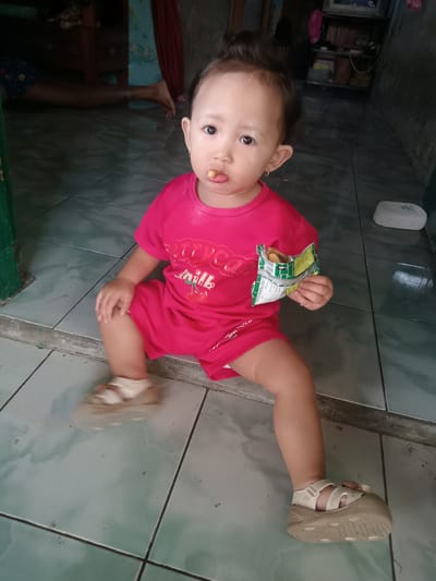 A toddler is sitting on a tiled floor, wearing a red shirt and shorts, and beige sandals. They have a snack in their mouth and hold a partially opened packet of chips in their right hand. In the background, there are hints of a living area with furniture and a person's legs visible. The scene appears to be in a home setting, consistent with the city of Bungur, Indonesia. The floor has large square tiles.