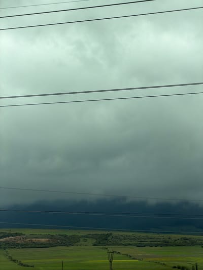The image shows a view in Mrgvali Chala, Georgia, with a landscape divided between sky and land. In the foreground, power lines stretch across the frame. The sky is overcast with grey clouds. On the right side of the image, a green landscape is visible, with trees and open fields. A power line tower is standing in the distance. A darkened area suggests a possible rain shower occurring in the middle ground. The image is taken from a low angle.