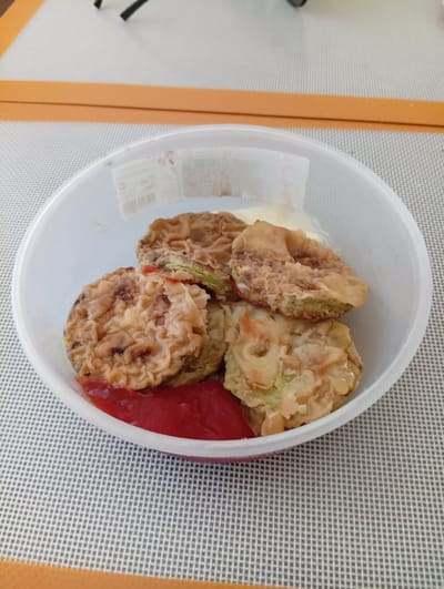 A close-up, overhead shot shows a white plastic container filled with fried food. The food appears to be sliced vegetables, possibly zucchini or eggplant, coated in a crispy batter and lightly fried. Some of the pieces are visibly green underneath the batter, confirming they are vegetables. A dollop of white creamy sauce, possibly mayonnaise or a yogurt-based dip, is visible at the top of the container. At the bottom of the container, there is a bright red sauce, likely ketchup. The container sits on a grey and white checkered placemat, which is on top of a beige surface. In the background, there are parts of a black object and a light orange border. The image was taken in Rezina, Moldova.