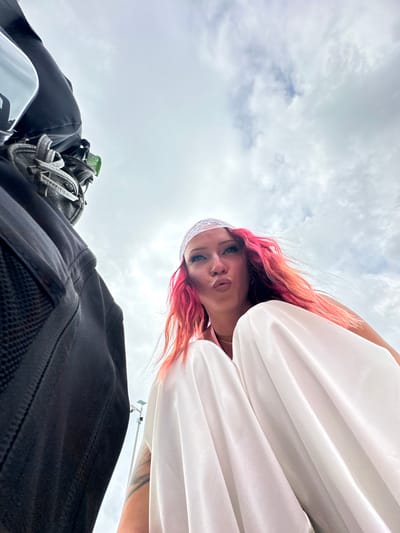 A low-angle shot shows a woman with pink hair, wearing a white top and headband, looking down at the camera. She has a nose piercing and light makeup. Behind her, a cloudy sky fills the background. In the foreground, a black bag or backpack is visible, along with some other items possibly strapped to it, including a water bottle. The location is unknown, but the prompt indicated the city of Kampung Toyapakeh, Indonesia. No specific actions or interactions are evident in the image.