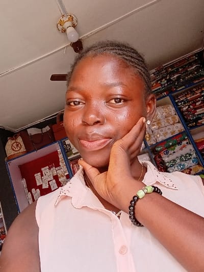 This image depicts a young adult female, appearing to be of African descent, positioned centrally in a close-up, selfie-style photograph taken indoors within a commercial establishment in Ayob, Nigeria. The female subject has dark, braided hair, a light brown complexion, and is looking directly at the camera with a slight smile. She wears a light peach/pink sleeveless collared blouse with a scalloped edge, a thin gold-colored necklace, a small stud earring in her right earlobe, and a beaded bracelet on her left wrist consisting of dark beads interspersed with two larger, light green, spherical beads. Her left hand is raised, with fingers gently resting against her chin and jawline.

The background reveals a retail environment consistent with a fabric or general merchandise store. To the subject's left (viewer's right), multiple shelves are stocked with neatly folded and rolled colorful fabrics featuring diverse patterns, some wrapped in clear plastic. To the subject's right (viewer's left), a multi-tiered display unit showcases various items: a light-colored decorative beaded object, a brown handbag with a "MK" logo, a red-backed display board presenting numerous small jewelry items (earrings, rings) attached to cards (some marked "24K"), and lower shelves with silver-toned accessories. The white ceiling features a decorative light fixture with a white energy-saving bulb and a partially visible dark ceiling fan.
