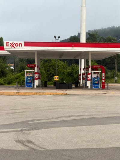 An exterior, medium shot captures an Exxon gas station located in Wildwood, United States. The scene is overcast with a foggy, mountainous backdrop. The gas station features a red and white canopy extending over gas pumps. Two blue and white Exxon Synergy gas pumps are visible on the left, while two red and white Synergy pumps are on the right. Trash cans are situated behind the pumps. A yellow sign with red lettering and the words "HONEST BROTHERS" is mounted on a pole to the right of the pumps. The asphalt in the foreground shows tire marks.