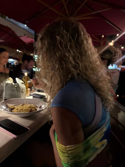 At an outdoor restaurant in Honolulu, United States of America, a person with long, curly blonde hair leans over a plate of pasta. The person wears a blue and green dress. On the table, a phone with a pink case sits next to the plate. Other people are in the background, including a person with a beard and a woman. Red umbrellas provide shade above the table.