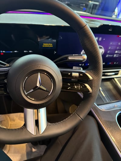 The image shows the interior of a car, focusing on the steering wheel and dashboard. The steering wheel is black leather with perforated grips and a Mercedes-Benz logo in the center. On the left side of the steering wheel, there are controls, including a silver volume dial labeled "VOL". The dashboard features two large digital screens. The screen on the left displays vehicle information such as speed (75 km/h), distance (461 km), and outside temperature (65.7, 108). A small yellow notification box on this screen reads "Modalità notturna parzialmente attiva" (Night mode partially active). The screen on the right displays a purple interface with icons and text, possibly navigation or infotainment options. Ambient lighting strips, a mix of pink and blue, are visible above the dashboard. The car appears to be parked or stationary. The presence of a Mercedes-Benz vehicle and its modern digital interface suggests a contemporary urban environment, possibly a dealership or a public parking area in Parma, Italy, given the context of the request. The lighting suggests it is daytime.