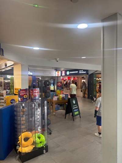 The image depicts an interior scene, likely within a shopping center or travel services hub, showing a brightly lit retail environment. In the foreground, a display case contains various travel pillows, one yellow and U-shaped and another green and somewhat irregularly shaped, possibly a character design. Adjacent to this is a blue rectangular structure. Further back, a shelf unit holds more pillows.
A prominent sign reads "Zone," suggesting a designated area. Signs for "KFC" and "Starbucks" are visible, indicating food and beverage services are available. A "Burger King" sign is also present. In the background, people are seen walking and interacting. One individual, wearing a light-colored t-shirt and patterned shorts, is walking away from the camera. Another person, wearing a grey t-shirt and blue shorts, stands facing away from the camera. A black and white dog is also visible walking on the floor.
The flooring is tiled in a neutral gray tone. Overhead, circular light fixtures illuminate the space, and a small green light is visible near the ceiling in the upper left corner. A security camera is mounted on the ceiling. The overall impression is one of a busy, functional retail space. The location is identified as Stroud, United Kingdom.