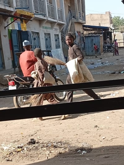 The image depicts an outdoor street scene in Kofar Ruwa, Nigeria, under clear daylight conditions. The ground is unpaved, consisting of packed dirt with scattered debris and litter. A prominent black horizontal bar partially obstructs the bottom portion of the frame.
In the midground, three primary figures are visible. To the left, a man wearing a white cap and a red-orange shirt is seated on a black motorcycle, facing away from the camera. Immediately to his right, a young individual dressed in a brown tunic and pants is positioned near the motorcycle, carrying a large bundle of stick brooms and a light-colored sack. Further right, another young individual, also in a dark brown tunic and pants with a dark cap, walks from left to right across the frame, carrying a large white sack draped over their left arm, and directly looking towards the camera.
The background features several structures. On the left, a multi-story concrete building with multiple blue-shuttered doorways and balconies with metal railings is visible. A sign partially reads "SANI ALUMANAH ENTERPRISES." To the right, smaller, possibly corrugated metal structures or kiosks line the street. Several other individuals are visible in the distance, including a child in a red shirt near a blue metal structure and adults near parked motorcycles. The overall environment appears urban with signs of commercial activity and unpaved thoroughfares.