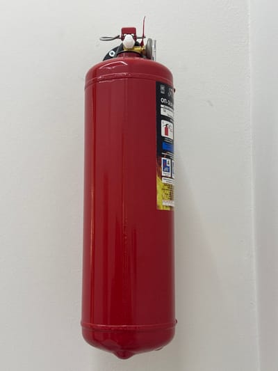 A red, horizontally-mounted fire extinguisher hangs on a white wall. The extinguisher has a pressure gauge, handle, and pin at one end. A label with instructions and symbols is wrapped around the cylinder. The cylinder has rounded ends and a glossy finish. The extinguisher appears to be securely mounted.