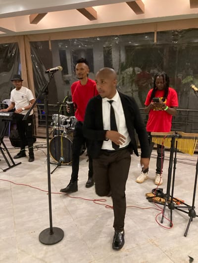 The scene depicts four males engaged in a musical performance or rehearsal within an indoor venue in Murera ward, Kenya. The foreground features a male of dark complexion, clean-shaven head, wearing a white collared shirt, black tie, black blazer, dark brown trousers, and black dress shoes. He is in mid-stride, right leg raised, left arm slightly extended, with an open mouth, indicative of singing or speaking energetically towards the viewer's left.

To his left, another male with a dark complexion, black hat, white t-shirt, black trousers, and black shoes operates a black electronic keyboard positioned on a stand. A black microphone stand with a light-colored microphone is directly in front of the keyboardist.

Behind the foreground subject and to the left, a third male with dreadlocks styled upwards, wearing a red t-shirt and black trousers with black boots, stands holding a black tumbler in his left hand, looking towards the right. A partial drum set, including a blue bass drum, is visible behind him.

To the far right, a fourth male with dreadlocks, wearing a red t-shirt featuring a yellow graphic and black trousers with white sneakers, stands looking down at a black smartphone held in both hands. A light-green wristband is on his right wrist, and an ankle bracelet is on his left ankle. To his left, a percussion instrument resembling chimes or a xylophone on a black stand is present.

The floor is composed of light-colored square tiles. Red electrical cables are visible running across the floor in multiple locations. The background features large transparent panels, possibly windows or plastic sheeting, revealing blurry green foliage and reflections of structures outside. The ceiling displays exposed light-colored beams.