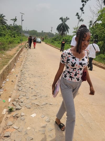The image depicts an outdoor scene on a sunny day along an unpaved road in what is identified as Candelaria, Colombia. The primary subject is a young woman, positioned in the foreground, walking from the right side of the frame towards the left. She has dark braided hair styled in an updo with a visible face-framing curl. She wears a white short-sleeved top adorned with multiple black Playboy bunny logos, grey sweatpants, and black slide sandals with a white "TF" logo. A gold-colored chain necklace with a small pendant is visible around her neck, and a maroon shoulder bag hangs from her right side. She carries a colorful spiral-bound notebook or planner in her left hand, looking downwards and slightly to her right.

The road surface is primarily dirt and gravel, with numerous broken concrete or asphalt fragments scattered, particularly along the left side. A concrete curb or drainage ditch runs parallel to the road on the left, containing some litter. Dense green tropical vegetation, including tall palm trees, lines both sides of the road under a hazy or overcast sky. Several streetlights, appearing to be solar-powered, are spaced along the road.

In the mid-ground, further along the road, several other individuals are present. On the left side of the road, two people walk towards the camera, one in dark clothing and another in a red top and dark pants. Further ahead, a light-colored car (possibly silver) is visible. On the right side of the road, two individuals walk away from the camera: one wears a black t-shirt with orange lettering and dark pants, and another wears a white t-shirt, dark pants, and carries a black shoulder bag. No direct interactions between the individuals are discernible.