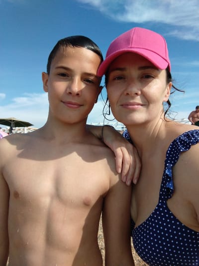 This image captures two individuals on a beach in Blagoveschenskaya, Russia, under a clear blue sky with sparse white clouds. In the foreground, a young male, appearing to be an adolescent, stands shirtless with visible water droplets on his skin. He has short, dark, wet hair and a faint smile, looking directly at the camera. His left arm extends around the right shoulder of the adjacent female. To his right, an adult female with a light complexion and visible freckles wears a bright pink baseball cap and a dark blue swimsuit top patterned with white polka dots and ruffled straps. Her dark hair is wet, with strands clinging to her face and neck. She also exhibits a faint smile and looks towards the camera. Her left forearm rests on the young male's left shoulder, and a silver-toned bracelet is visible on her left wrist. Both individuals appear wet, suggesting recent contact with water. In the mid-ground, a dark green beach umbrella is visible on the left, alongside a white structural element. A partially obscured, shirtless male figure is seen in the far right background, walking away. The ground in the lower right foreground consists of coarse sand or pebbles.