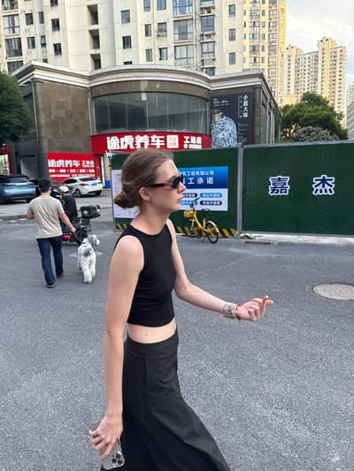 The image, rotated, depicts an urban street scene in what appears to be Putuo District, China, during daylight hours. The primary subject is a young woman standing in profile, wearing a black sleeveless top, black pants, and dark sunglasses. She holds a smartphone in her left hand and makes a finger heart gesture with her right hand, looking upwards. Her hair is pulled back.

In the background, another individual, a man, is seen walking a white, fluffy dog on a leash. A yellow shared bicycle is parked nearby. The street is lined with various buildings, including residential structures with numerous windows and visible air conditioning units, and commercial storefronts.

Prominently displayed is a red sign for "途虎养车" (Tuhu Car Maintenance) with the text "TUHU.ON" and details like "保养维修: 洗车美容" (Maintenance repair: car wash beauty), "上海中两城店: 18517553303" (Shanghai Zhongliangcheng Store: phone number), and "途虎养车工场店" (Tuhu Car Maintenance Workshop Store) with the number "952688".

Further along the street, a dark poster advertises "小器大琢" (A small item, a big work/carving), likely for an exhibition. Adjacent to this, a green construction barrier features large characters "嘉杰" (Jiajie) and a blue sign stating "筑工程有限公司" (Construction Engineering Co., Ltd.). Below this, "工承诺" (Worker's Promise) lists four points: "运不扰民" (Transportation does not disturb residents), "配給不沾毛" (materials not shedding/dusting, as per OCR), "扬尘不超标" (Dust does not exceed standards), and "作业不违章" (Operations do not violate regulations).

The scene conveys a bustling city environment with ongoing commercial and construction activities. The woman's gesture and upward gaze suggest a relaxed and perhaps playful mood.