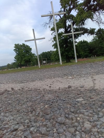 The image shows a gravel road in Stilwell, United States, leading to a cemetery. Three white crosses stand at the entrance to the cemetery, with trees and greenery visible in the background. The sky is overcast. The ground in the foreground is covered in small rocks and pebbles. A fence is visible in the background, marking the perimeter of the cemetery. Tombstones and grave markers are scattered within the cemetery grounds.
