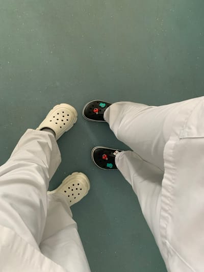 An overhead view captures the lower extremities of two individuals standing on a speckled, light blue-green linoleum floor, consistent with an institutional or medical facility within Nürtingen, Germany. Both individuals wear white uniform trousers. The person positioned on the left wears a pair of cream-colored, perforated clogs with thick, ridged soles. The person on the right wears a pair of black, perforated clogs. Each black clog is adorned with two distinct decorative charms: a red stethoscope and a light blue medical bag or fish-shaped emblem. The feet are positioned in close proximity, suggesting the individuals are standing side-by-side.