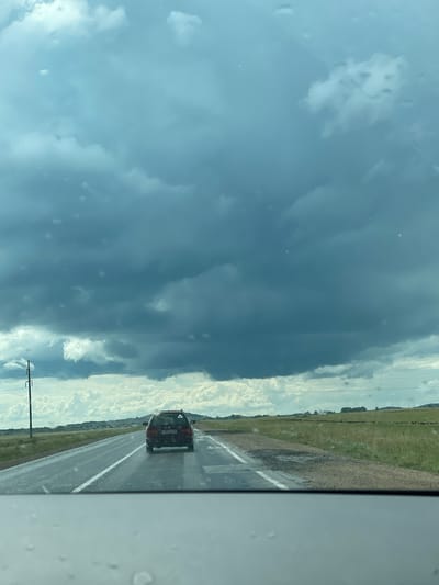 A dark, overcast sky with heavy, bruised-looking clouds dominates the upper portion of the image. Light rain or mist is falling, evidenced by water droplets on the windshield in the foreground and the wet appearance of the asphalt road. A red station wagon, with license plate 787 A36 15, is driving away from the viewer on the right lane of the road. The road is flanked by grassy fields. In the distance, a line of trees and some low hills are visible, with power lines and a utility pole to the left of the road. The scene appears to be a rural setting in Kazakhstan, possibly near the city of Жаңаауыл.