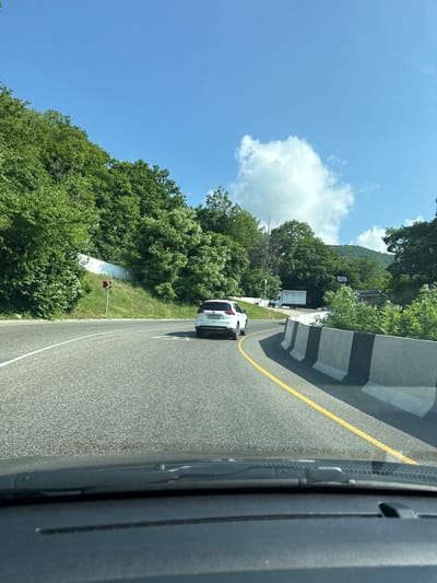 A white SUV is driving on a road with a yellow line. The road is surrounded by trees and hills, and there is a blue sky with white clouds. The image appears to be taken from inside another vehicle. The location is identified as Dederkoi, Russia.