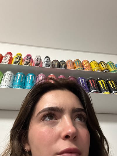 A person's face fills the right side of the frame, looking slightly upward. To the left, a tall shelf is densely packed with energy drink cans. Brands like Monster and Prime are recognizable in a variety of colors and flavors. The background is a plain white wall, providing a stark contrast to the vibrant display of cans. The focus is on the person's face and the adjacent collection of energy drinks.