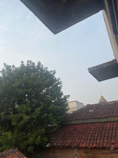 The image shows a view from a building's upper floor in Pasir Gadung, Indonesia, looking out over a residential area. In the foreground, the edge of a concrete structure juts out from the top right and top left, framing a hazy sky. To the left, a large, dense green tree dominates the frame. Below the tree, red tiled roofs of houses are visible, with one house featuring a distinctive yellow and green dome, possibly a mosque. The sky is overcast and pale, suggesting it is either early morning or late afternoon, or potentially a day with poor air quality. The scene captures a typical urban residential landscape in Indonesia, with a mix of vegetation and buildings.