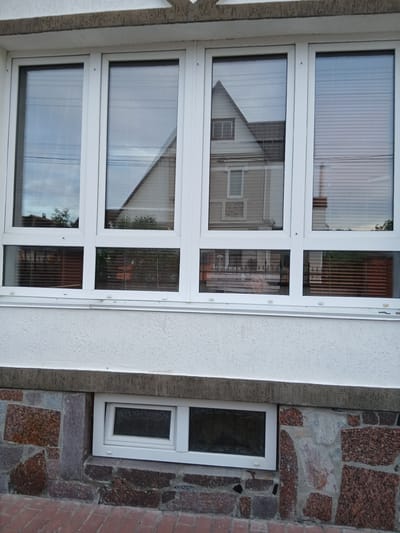 The image shows the exterior of a building in Vita-Poshtova, Ukraine. The building features large white framed windows, some of which are covered with horizontal blinds. The upper part of the building is textured white stucco. Below the large windows, there is a stone foundation constructed with irregular pieces of granite and other dark-colored stones. Further down, there is a small, horizontally oriented white-framed window with frosted glass. The ground in front of the building is paved with red bricks laid in a cobblestone pattern. Reflections in the windows show houses and sky.