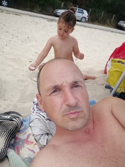 In the city of Osenovo, Bulgaria, on a sandy beach, a man is taking a selfie. In the foreground, the man's face, chest, and shoulder are visible. He is lying down, with his head turned towards the camera. Next to him, a colorful beach towel and a pair of black Adidas sandals are placed on the sand. In the background, a young girl is playing on the sand, holding a small seashell. Behind her, two cars are parked. The environment is a typical beach scene with sand and some scattered debris.