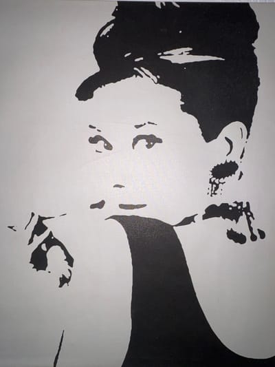 This is a black and white stencil art piece depicting the face of a woman. The style is minimalistic, with bold black shapes against a lighter background, suggesting a famous cultural icon. The subject's eyes are looking slightly to the left, and her hand is raised to her mouth, as if holding something. The overall impression is one of elegance and contemplation. The artwork is displayed in a public space in Aradippou, Cyprus, possibly on a wall or facade, contributing to the urban landscape of the city. The exact location within Aradippou is not specified, but its presence indicates an appreciation for art and public expression in the community.