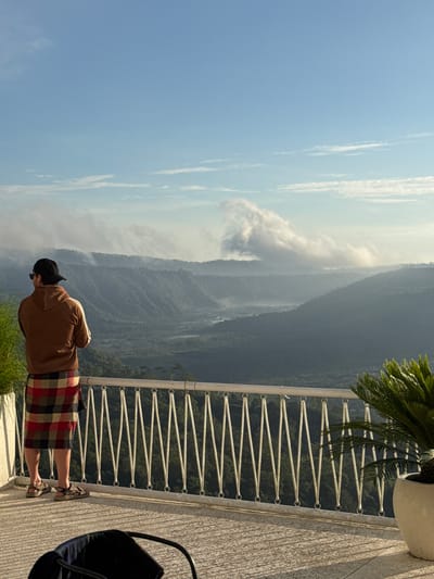 A full-body view captures an individual on a balcony overlooking a mountainous landscape. The person is wearing a brown jacket, a plaid skirt, sandals, and a black baseball cap. The balcony features a white railing, and plants in a white pot. In the background, a large mountain range extends under a hazy blue sky. The photo suggests the location might be in Kedisan, Indonesia, given the landscape features.