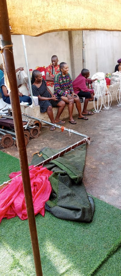 In Udi, Nigeria, an outdoor scene unfolds under a makeshift yellow-beige tarp canopy. The ground surface consists of a concrete slab, with sections covered by artificial green turf. In the foreground, a tall, weathered wooden pole stands vertically. To its left, a bright pink fabric lies crumpled on the turf, adjacent to a folded dark green, coarse fabric. Immediately behind these fabrics, on the concrete, a pile of rusted metal pipes and circular metal components are visible, alongside an L-shaped metal frame wrapped in white material.
In the midground, five individuals are seated. On the left, partially obscured, a person in a dark blue garment is visible. To their right, a woman with braided hair, wearing a black dress with white polka dots, sits on a low concrete ledge, looking towards her right. Next to her, a man with short hair, dressed in a red, orange, and green patterned shirt, sits on a red cushion, looking downwards. Adjacent to him, a woman with braided hair, wearing a multi-colored patterned top (green, red, yellow, black) and dark shorts, is seated on the concrete ledge. She is smiling, has a camera strap around her neck, and is looking towards her left. To her right, a young male or adolescent in a maroon hoodie and dark shorts sits on the ledge, extending his right arm towards white, decorative wireframe objects on the ground. A white bag, partially labeled "GOLDEN PENNY PURE FLOUR," rests near these objects.
The background features a light-colored building wall. The people appear to be engaged in conversation or resting in an informal setting.