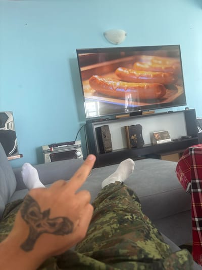In an indoor living room setting, a person is captured from a low-angle perspective, lying on a gray sofa. The person is wearing white socks and camouflage-patterned pants. Their right hand is raised, with the middle finger extended, and a skull tattoo is visible on their hand. The room's ambiance is illuminated by natural light, casting a soft glow across the scene. A television is mounted on the wall, displaying an image of sausages cooking in a tray. Below the television, a black shelf unit holds several items, including a stereo system, decorative objects, and a calendar. A red and black plaid blanket is draped over the right side of the sofa. The walls are painted a light blue, and a wall sconce is attached to the upper left portion of the wall. There are no visible indications of the city of Simeonovo, Bulgaria.