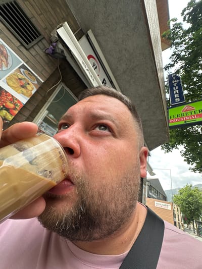 A man is depicted in a close-up, tilted selfie, drinking from a clear glass containing a light brown, foamy beverage, likely an iced coffee. He has a short beard, light-colored eyes, and his hair is dark with some graying at the temples. He wears a light pink t-shirt and a dark strap across his shoulder, possibly from a bag. His gaze is directed towards the camera.
The scene is outdoors in an urban setting under an overcast sky. In the immediate background to the left, parts of a building with brick patterns and a metal vent are visible, along with multiple picture boards displaying various food items, such as pancakes and other dishes.
Further in the background, several signs are discernible. A blue sign, oriented vertically, partially reads "PINTURES" (Paints). Adjacent to it, a green sign, also vertical, states "FERRETER INDUSTRIAL," indicating a hardware store, and includes an arrow pointing right. In the distance, another building features a sign with "werzalit" and "EXKLUSIV-HOLZMODULEN" (Exclusive Wood Modules) written on it. The text on the signs suggests a Spanish or Catalan-speaking region, aligning with the provided location of Andorra la Vella, Andorra. The activity appears to be a casual moment of refreshment outdoors.