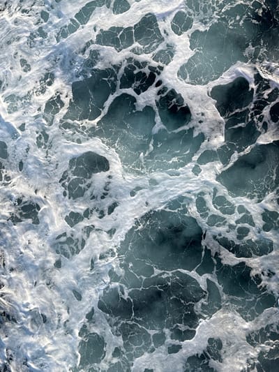 This image displays an overhead, high-angle view of turbulent sea water in the marine environment near Kamariotissa, Greece. The primary subject is the dynamic interaction between white foam and the underlying blue-green water. Irregular, fractal-like patterns of bright white foam cover large portions of the water surface, forming interconnected networks and isolated frothy patches. These patterns suggest significant surface agitation, indicative of waves, currents, or a wake. The water itself varies in tone from a deep, darker blue-green in calmer or deeper apparent sections to a lighter, more vibrant hue where the foam is thinner or sunlight penetrates. No people, vessels, or specific man-made objects are present within the frame. The scene is exclusively natural and aquatic.