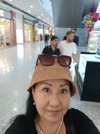 In Kırıkkale, Turkey, a medium shot captures a woman in the foreground wearing a straw hat and sunglasses, her face angled towards the camera. She has dark hair and is adorned with pearl earrings and a pearl necklace. Behind her, out of focus, two men are visible in a brightly lit indoor environment, possibly a shopping mall. One man wears a white t-shirt, and the other a dark polo shirt and a baseball cap. A display of colorful balloons is also present in the background. The environment appears to be a commercial space with shops and a polished floor.